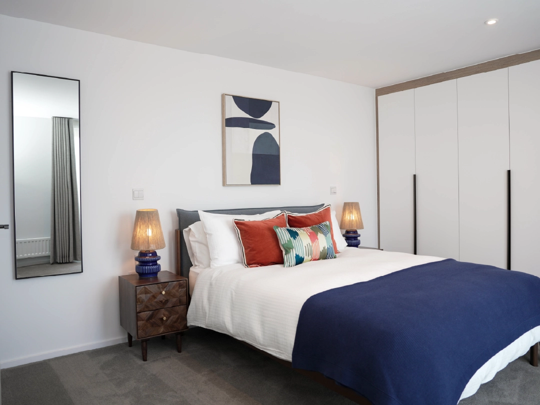 Large master bedroom, with navy double bed and accent cushions, dark wooden side tables and fitted wardrobes in a home at Brabazon, Bristol. 