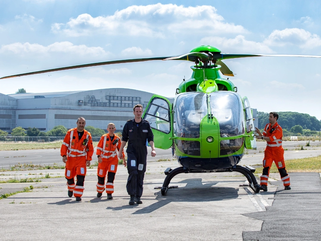 A bright green air ambulance helicopter on a tarmac, with four crew members in orange and black uniforms walking towards the camera. The background features an industrial hangar and a partly cloudy sky.