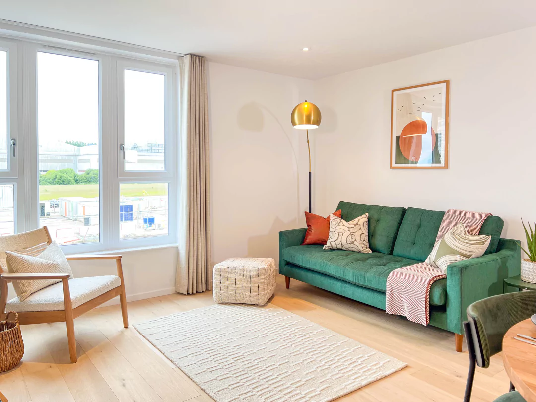 Light and spacious apartment living room, with a cream rug, modern green couch with assorted pillows, single arm chair and a brass gold free standing lamp. 
