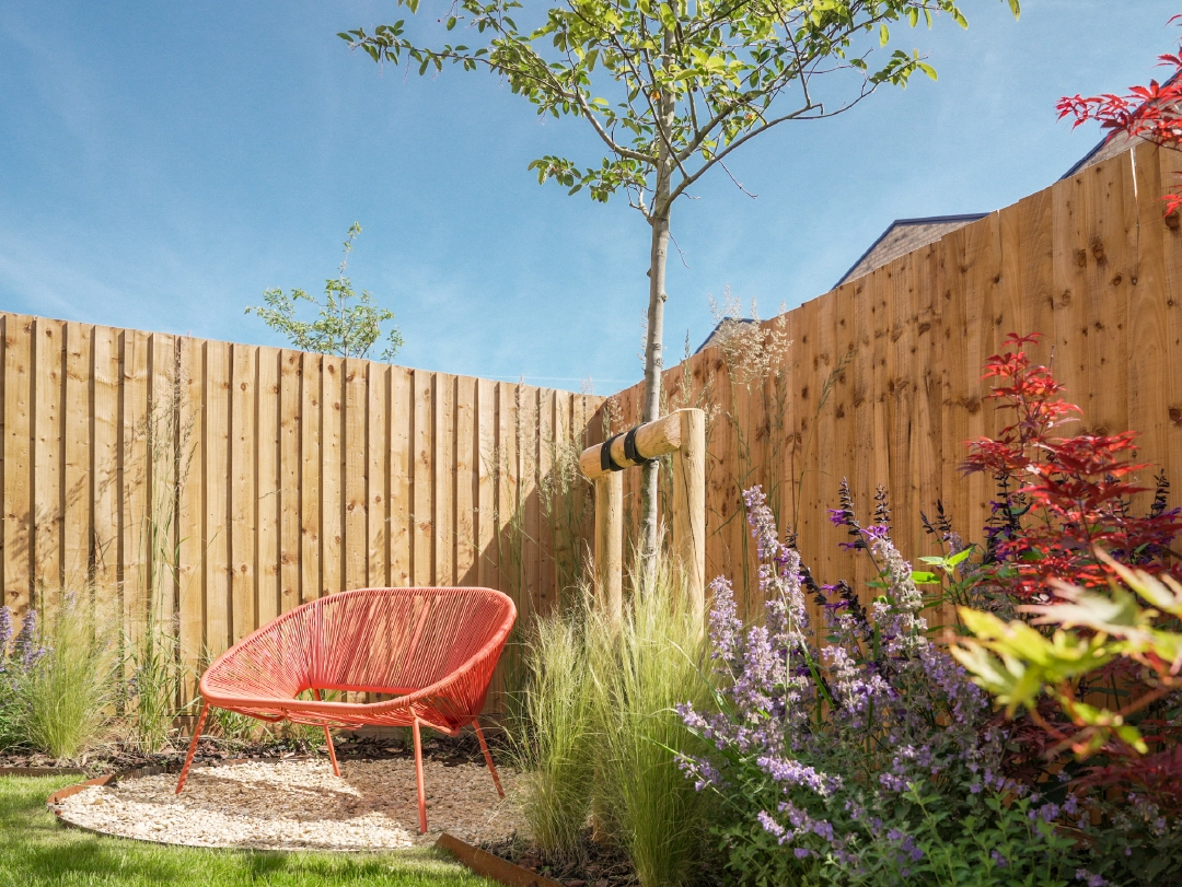 A vibrant red chair sits in a sunny garden corner at The Coanda at Brabazon, surrounded by spring flowers and a wooden fence under a clear blue sky.